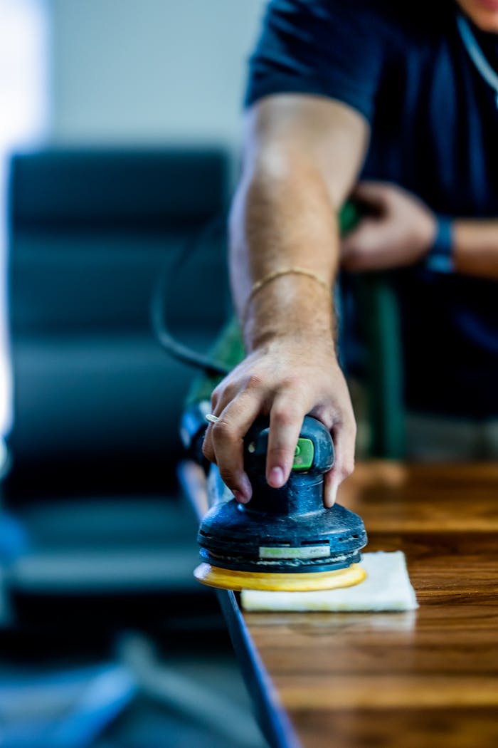 A close-up of a man's hand using a buffing machine on a wooden table indoors.